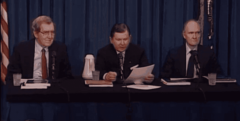 Three old white men wearing suits and sitting at a table. They all have microphones in front of them and are reading from prepared remarks